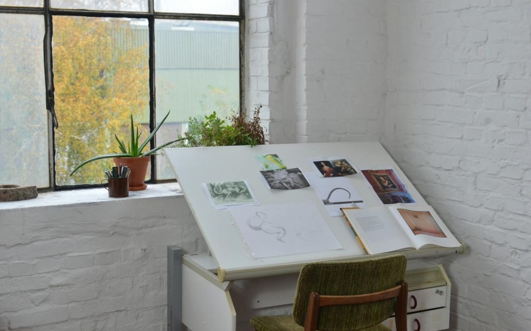 Opened books with various pictures and sketch placed on drawing table near window in workshop