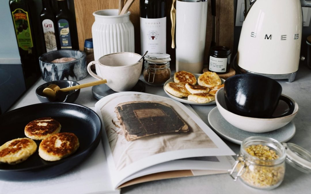 One portion of homemade cheese pancakes in frying pan and another on plate locating near opened stylish magazine among kitchen utensils and appliances in bright kitchen at home